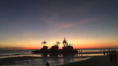 A beach at sunset with rocks and palm trees silhouetted against the sky.