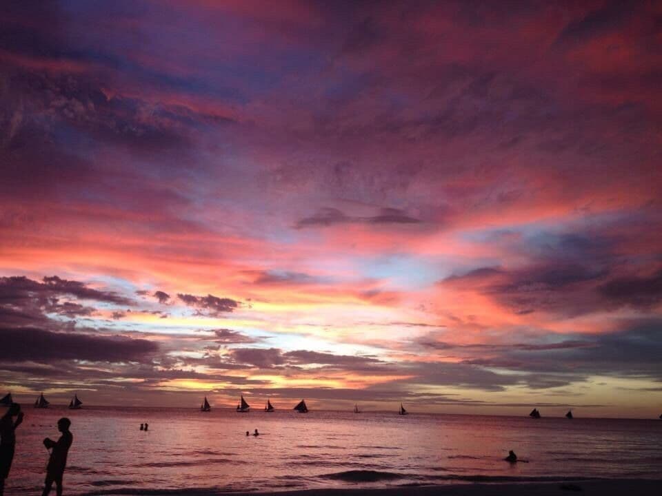 A sunset over a calm sea with sailboats and silhouettes of people.