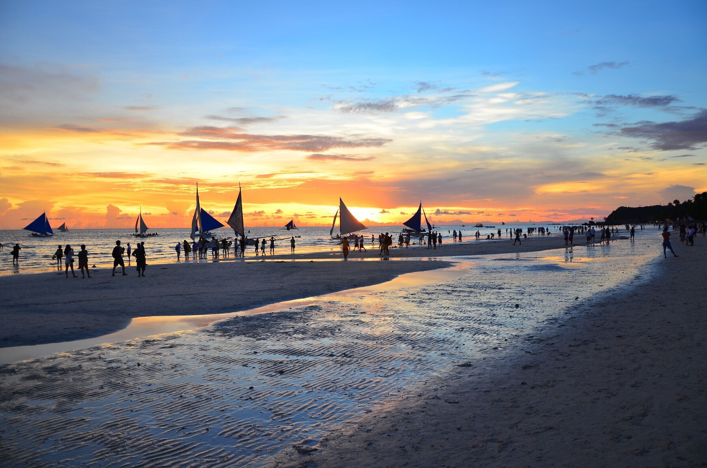 A beach at sunset with sailboats and people walking along the shore.