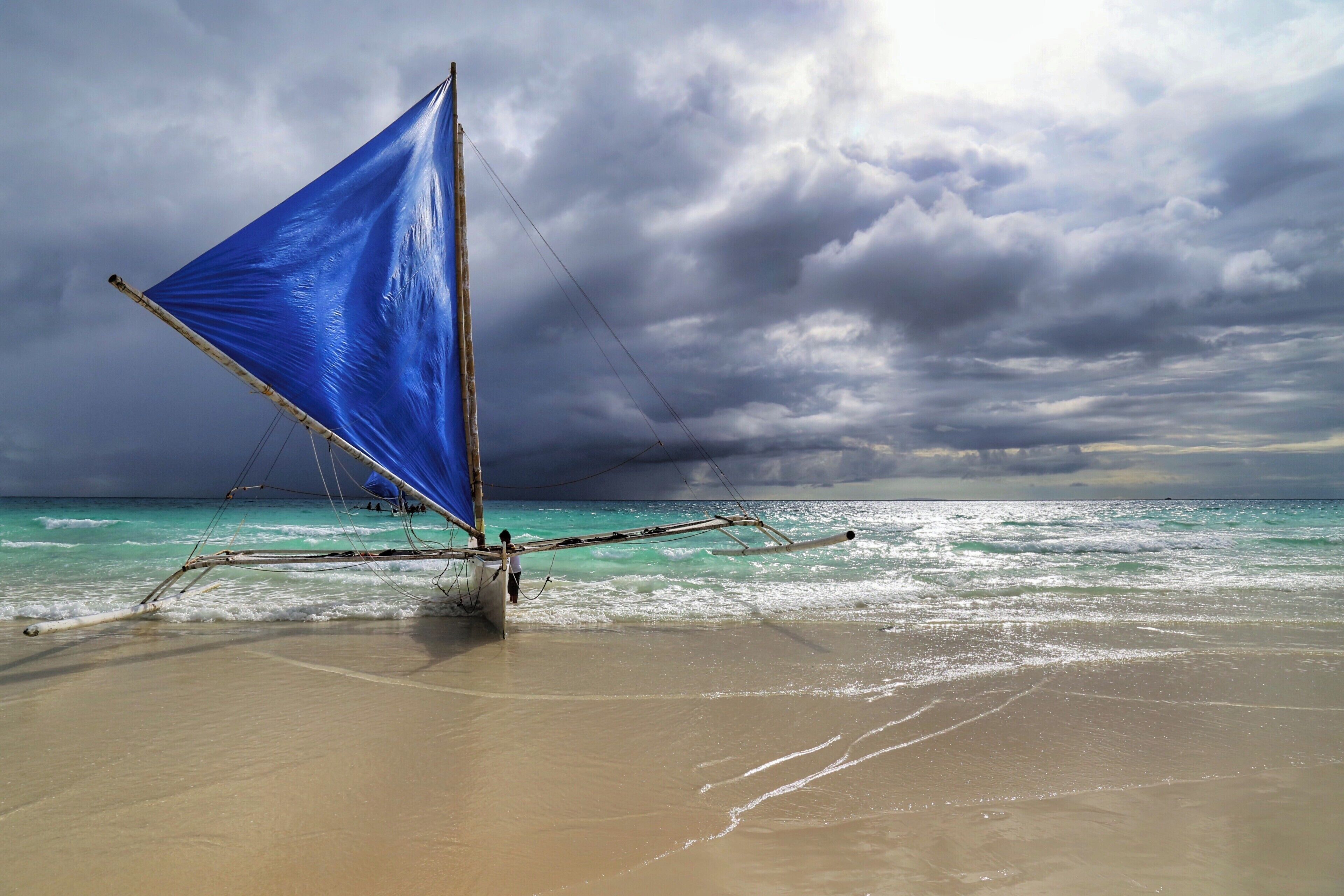 A sailboat with a blue sail on a beach with turquoise water and a cloudy sky.