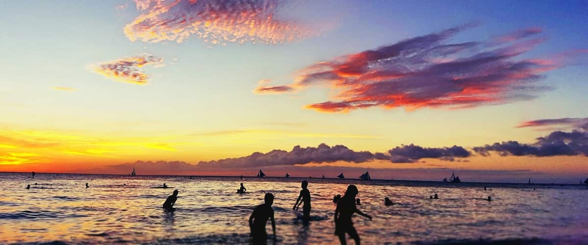 Atardecer sobre una playa con gente nadando y veleros a lo lejos.