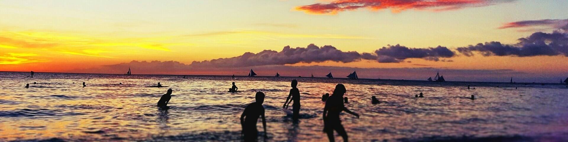 A sunset over a beach with people swimming and sailboats in the distance.