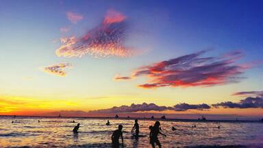 Atardecer sobre una playa con gente nadando y veleros a lo lejos.