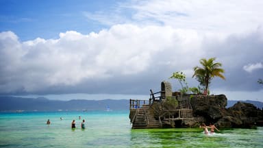 A tropical beach with clear turquoise water, a wooden staircase leading to a rock platform, palm trees, and distant mountains.