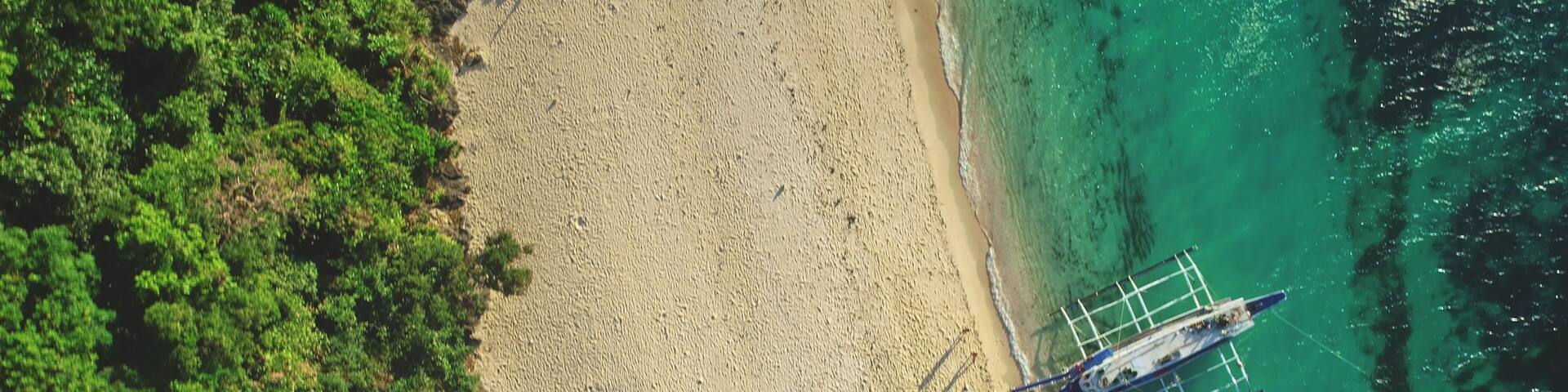 Aerial drone view of a tropical beach with turquoise water and white sand meeting a lush green forest, with a traditional outrigger boat anchored near the shore