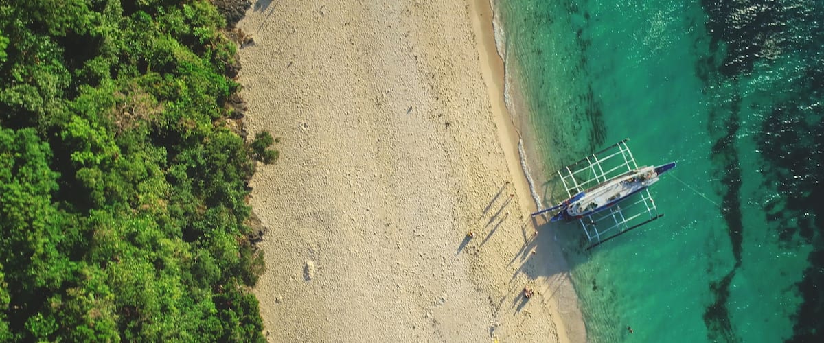 An aerial view of a beach with clear turquoise water, a sandy shore, and a boat anchored near the water's edge.