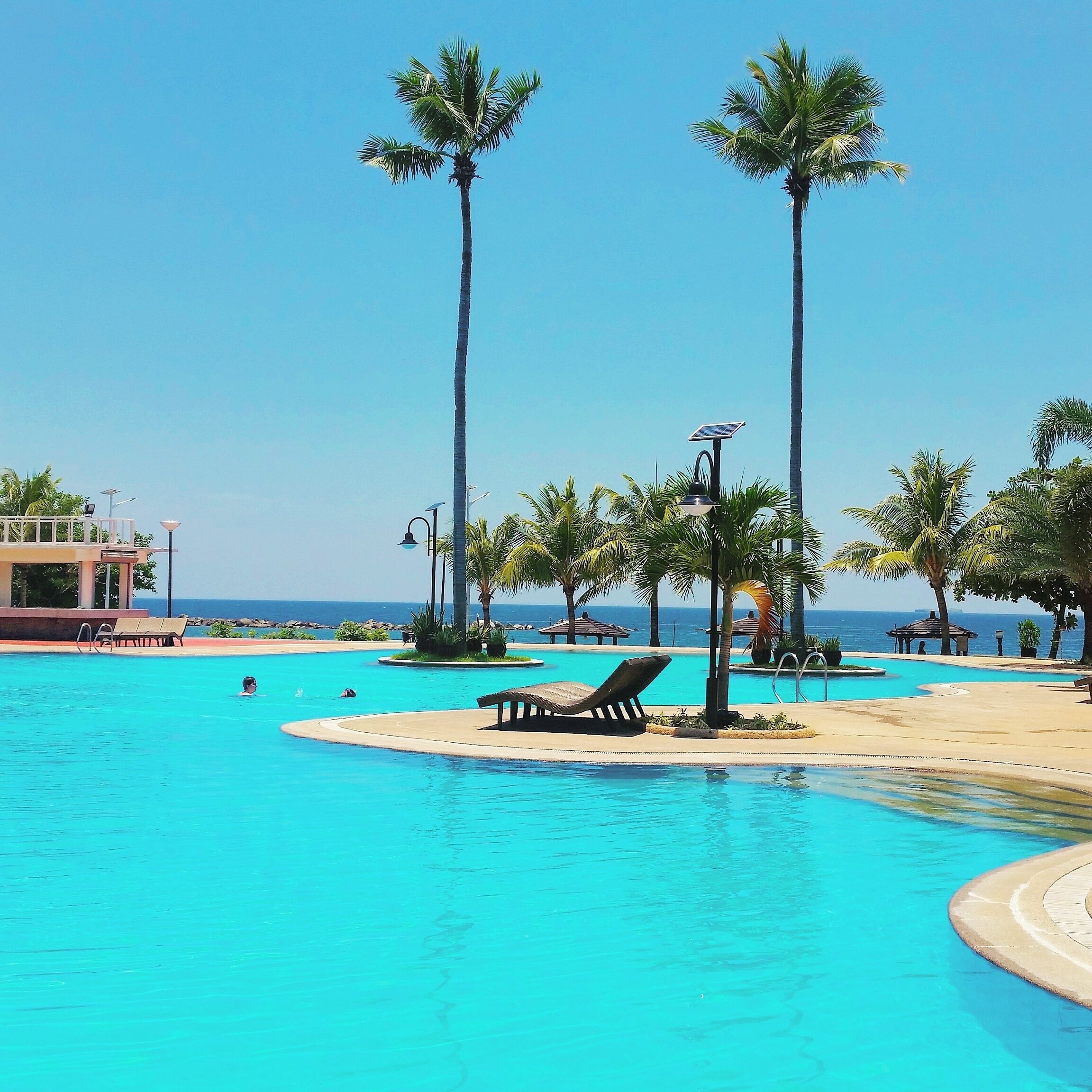 Piscina azul límpida com palmeiras, espreguiçadeiras e vista para o oceano.