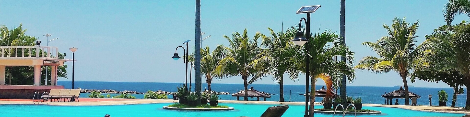 Piscina azul límpida com palmeiras, espreguiçadeiras e vista para o oceano.