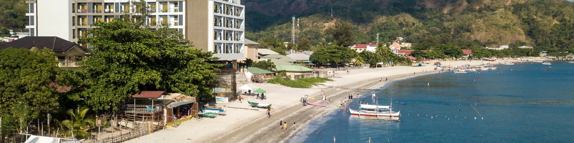 Una playa con barcos, un hotel y una colina boscosa al fondo.