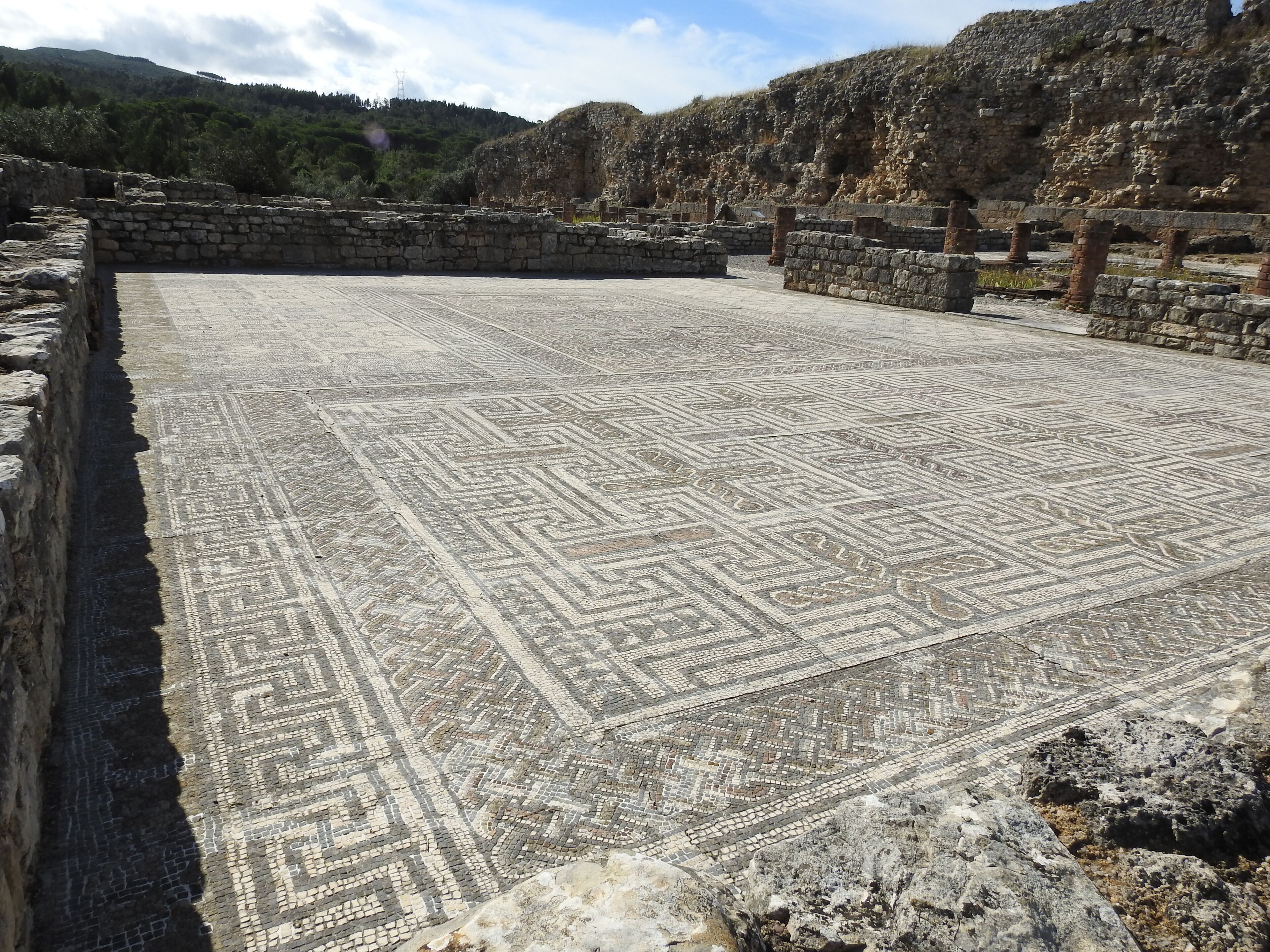 An ancient mosaic floor with geometric patterns, surrounded by stone walls and ruins.