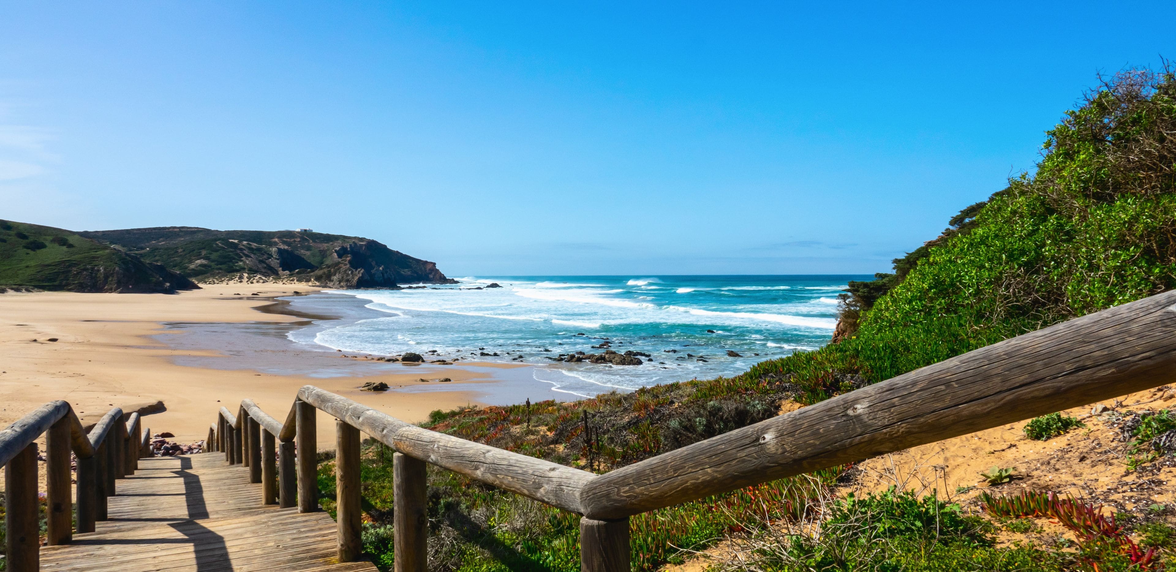 Eine Holztreppe führt zu einem Strand, wo Wellen an die Küste brechen, mit sandigem Strand und grünen Hügeln im Hintergrund.