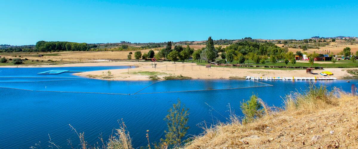 A lake with clear blue water, surrounded by dry grassland and trees.