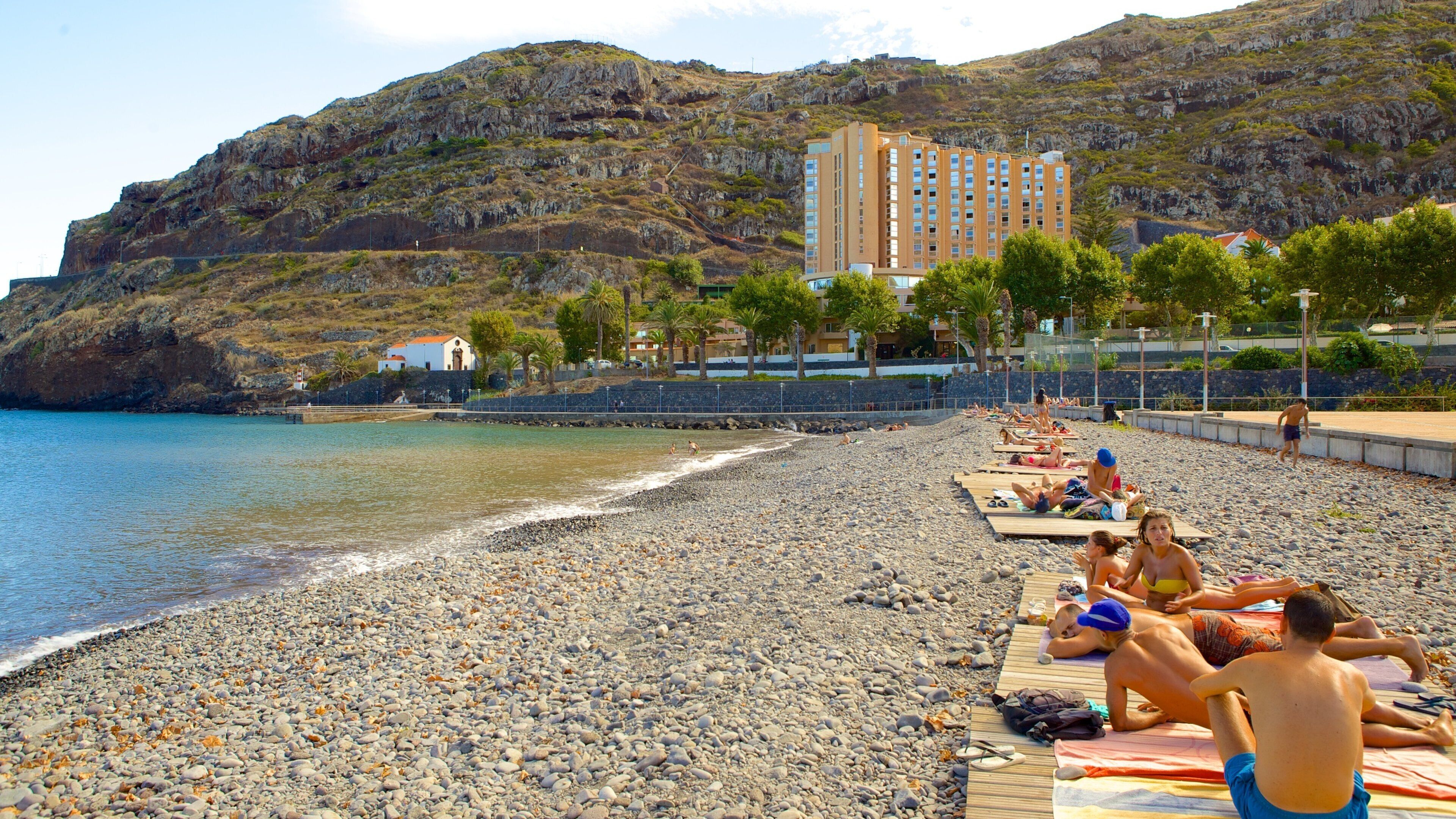 A pebbled beach with people sunbathing, a mountain in the background, and a building with a red roof.