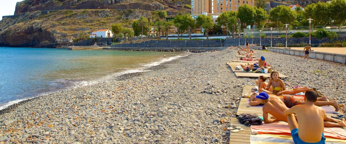 A pebbled beach with people sunbathing, a mountain in the background, and a building with a red roof.
