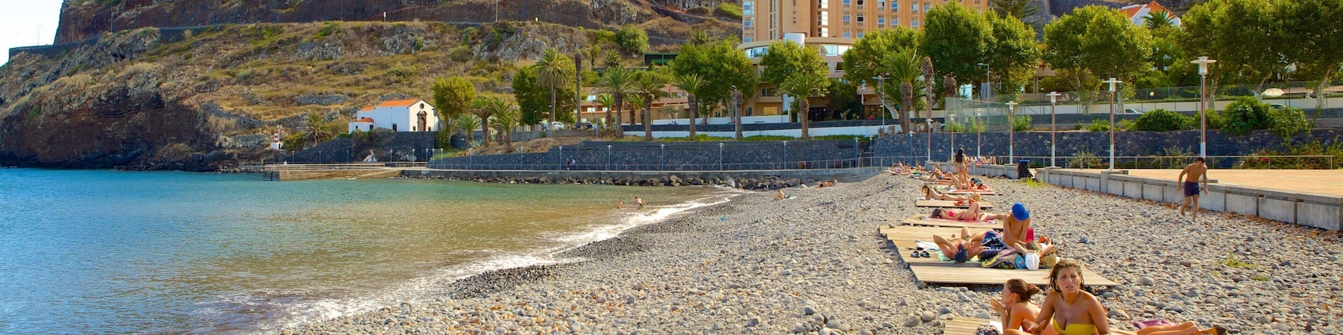 A pebbled beach with people sunbathing, a mountain in the background, and a building with a red roof.