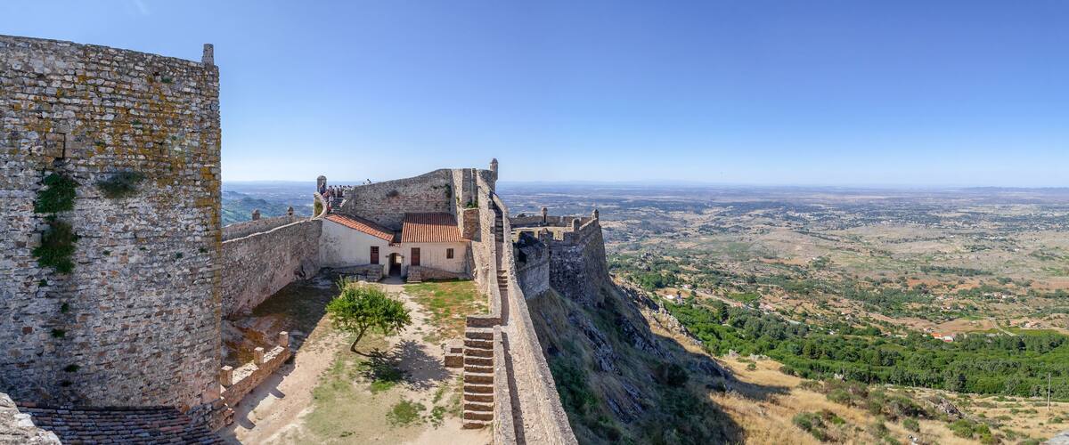 A historic stone wall with a view of a valley and a small building with a red roof.