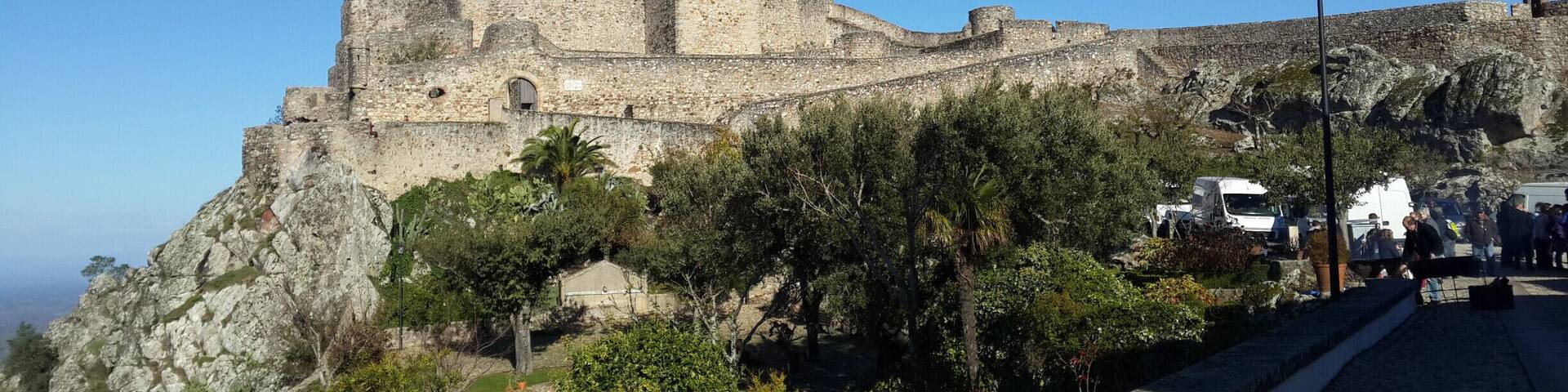 A historic stone fortress with a prominent tower, surrounded by lush greenery and a clear blue sky.