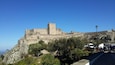 A historic stone fortress with a prominent tower, surrounded by lush greenery and a clear blue sky.
