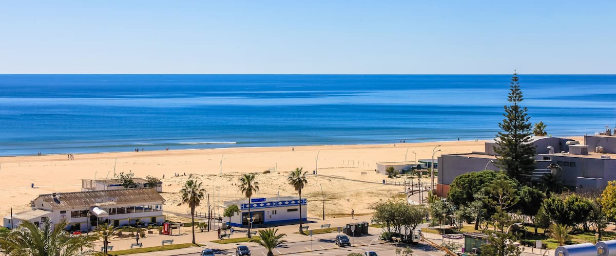 A beach with clear blue water, a sandy shore, and buildings in the foreground.