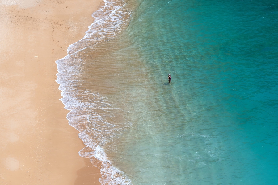 Una playa arenosa con agua turquesa transparente y una persona de pie en el agua.