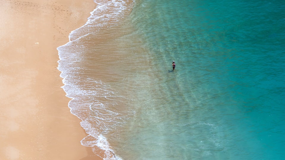 A sandy beach with clear turquoise water and a single person standing in the water.