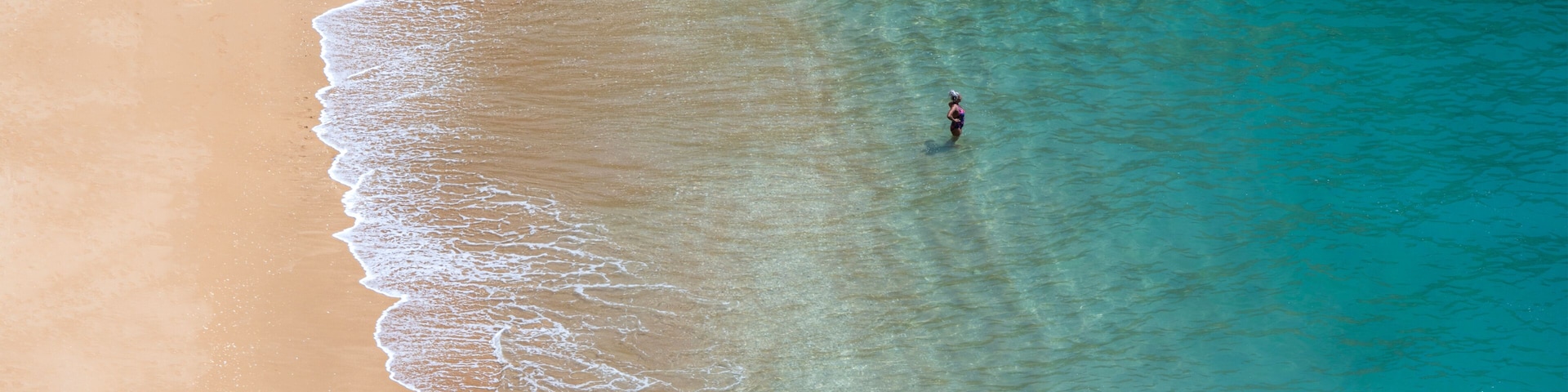 A sandy beach with clear turquoise water and a single person standing in the water.