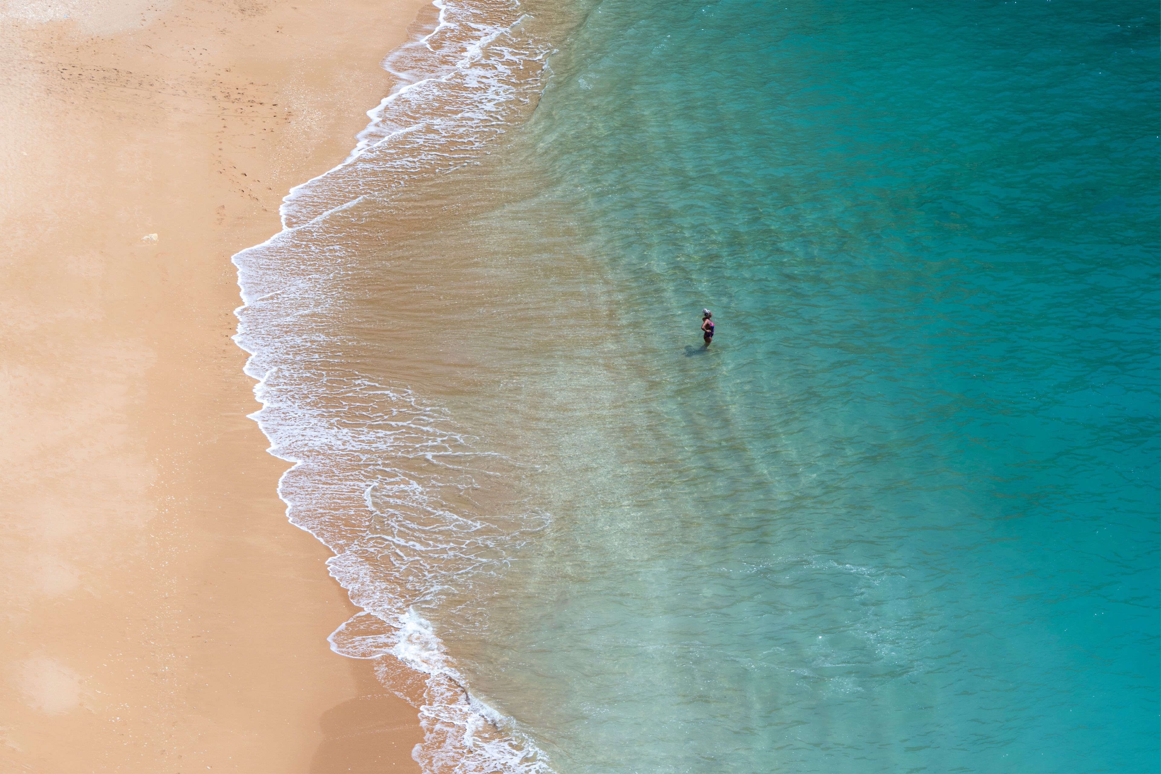 A sandy beach with clear turquoise water and a single person standing in the water.