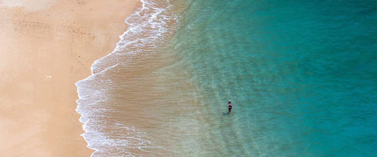 A sandy beach with clear turquoise water and a single person standing in the water.