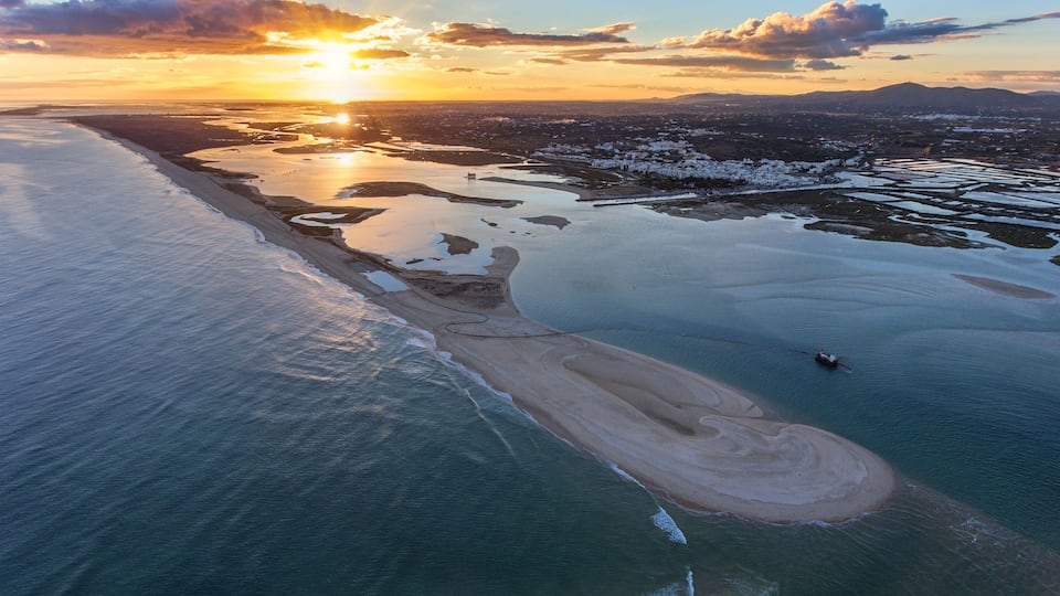 An aerial view of a coastal area with a beach, a boat, and a cityscape in the background during sunset.