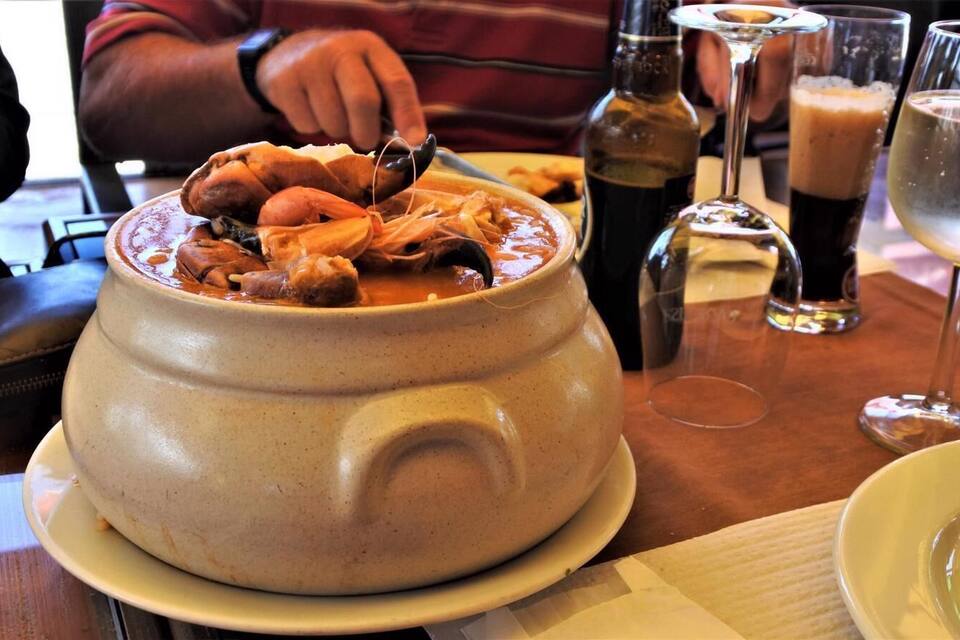 Una olla de cerámica grande llena de mariscos, sobre una mesa con vasos y botellas.