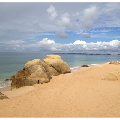 Una playa arenosa con grandes formaciones rocosas y un cielo despejado con nubes.