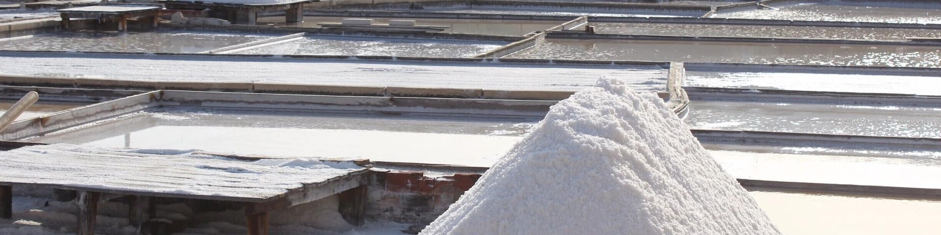 A salt evaporation pond with large white salt piles and a backdrop of houses and hills.