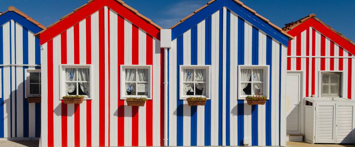 A row of colorful striped beach huts with white doors and windows, under a clear blue sky.