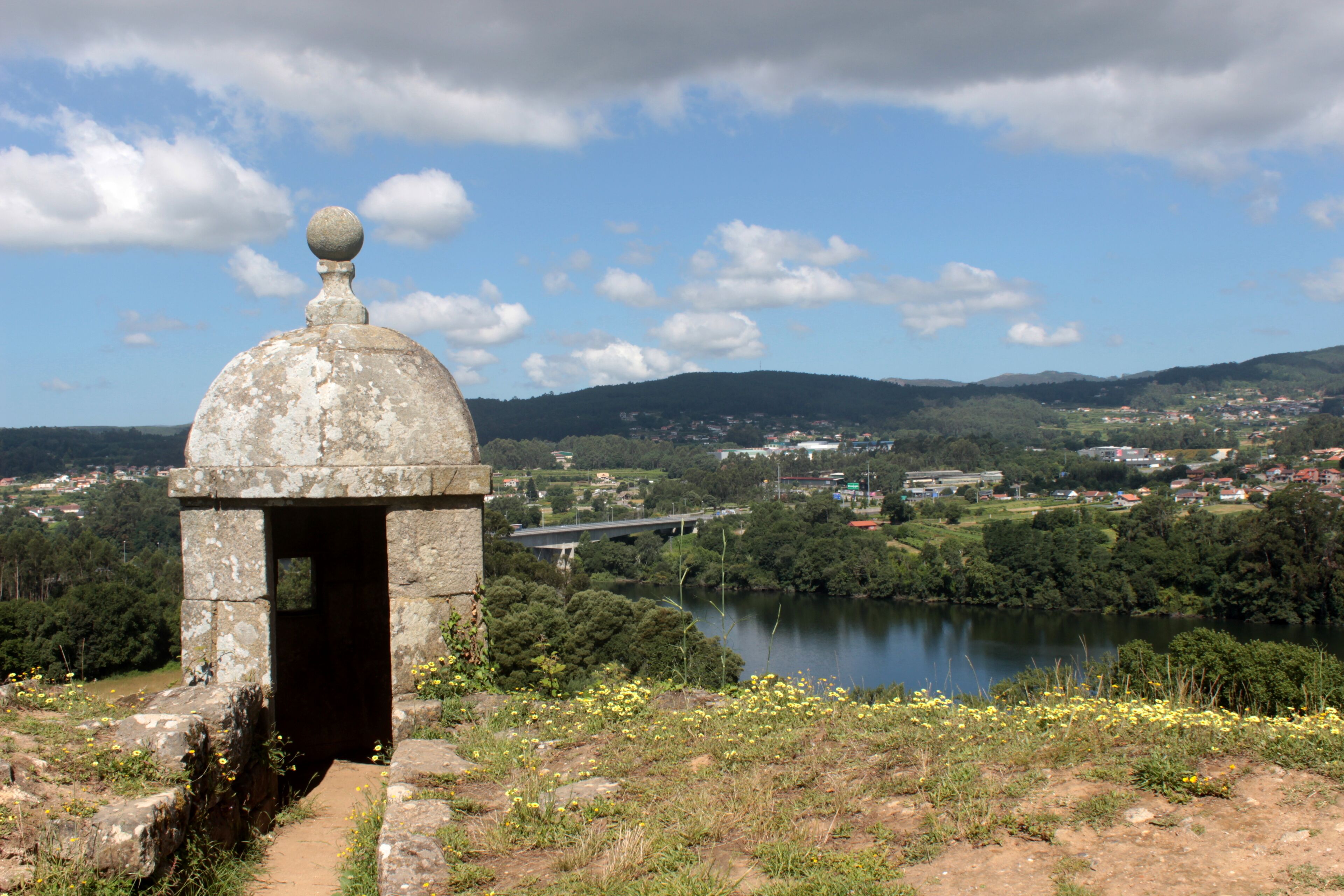 A stone structure with a dome and a ball on top, overlooking a river and a bridge in a hilly landscape.