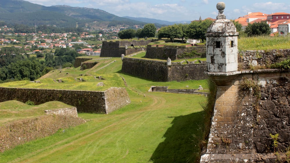 A historic fortification with stone walls and a watchtower overlooking a town and rolling hills.