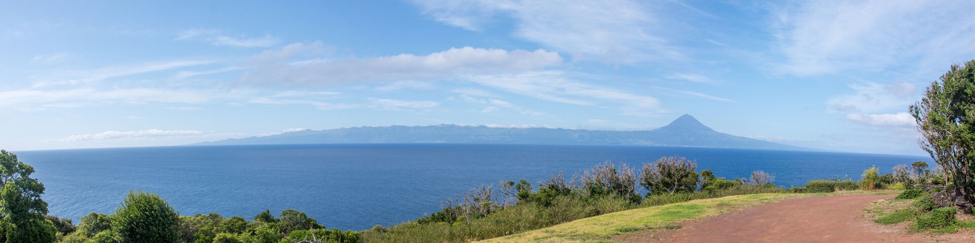 A coastal landscape with a dirt path, lush greenery, and a mountain in the distance.