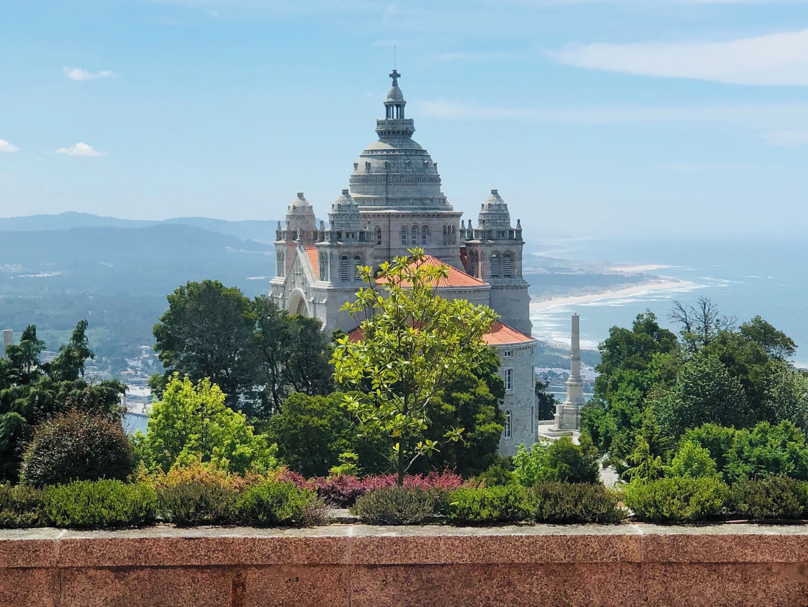 A historic cathedral with a red-tiled roof, surrounded by lush greenery and overlooking a coastal landscape.