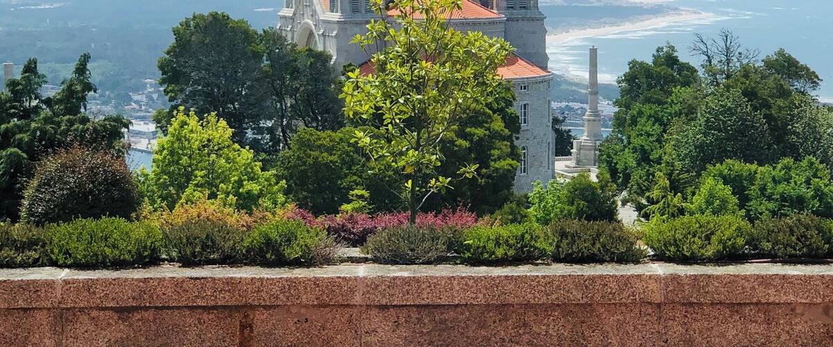 A historic cathedral with a red-tiled roof, surrounded by lush greenery and overlooking a coastal landscape.