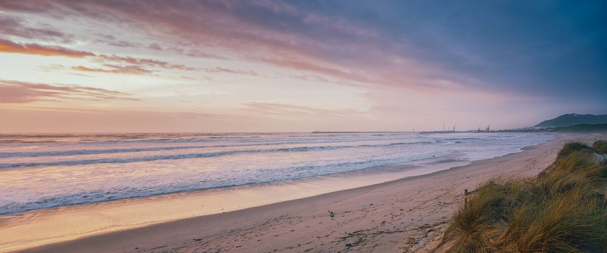A beach with gentle waves, a clear sky, and distant industrial structures.