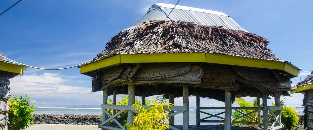 A thatched-roof gazebo with a wooden railing, surrounded by lush greenery and a clear blue sky.