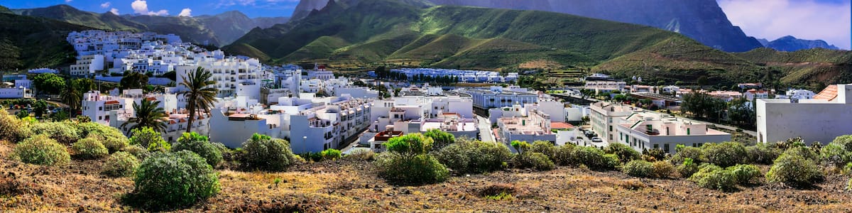 A coastal town with white buildings, lush green hills, and a mountain range in the background.
