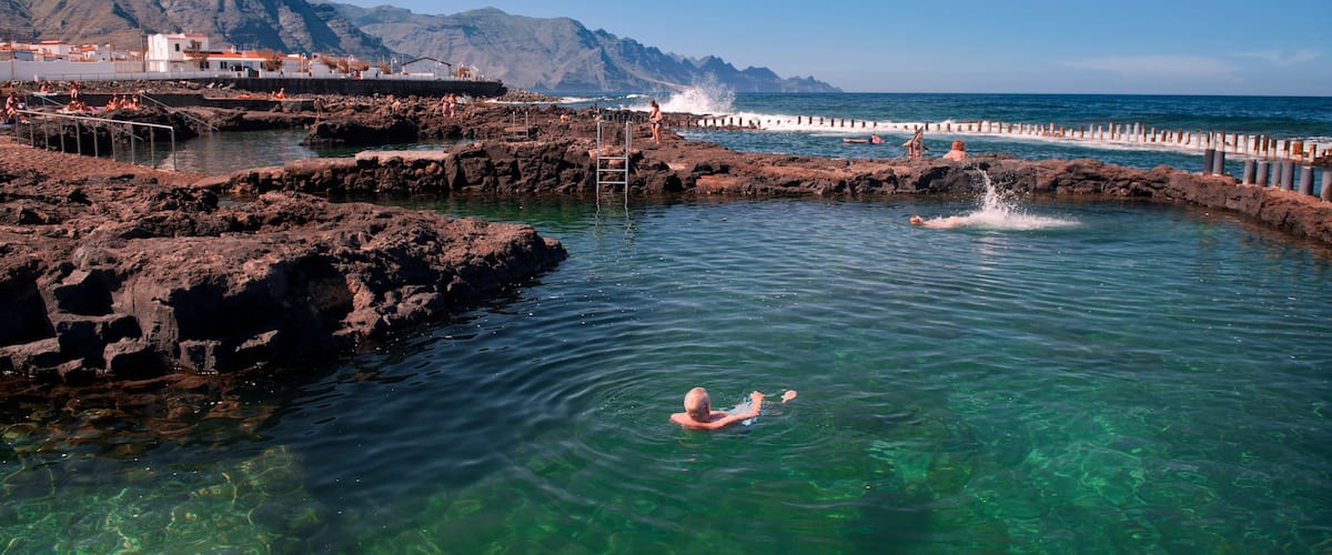 A clear blue sea with rocky formations, a person swimming, and a mountain in the background.