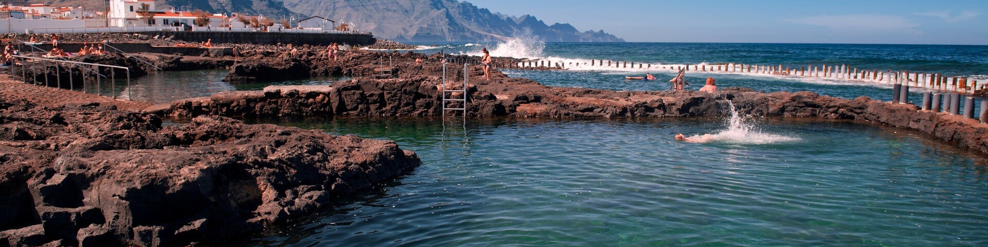 A clear blue sea with rocky formations, a person swimming, and a mountain in the background.