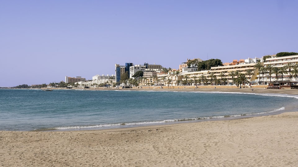 A beach with a clear sky, calm sea, and buildings along the shore.
