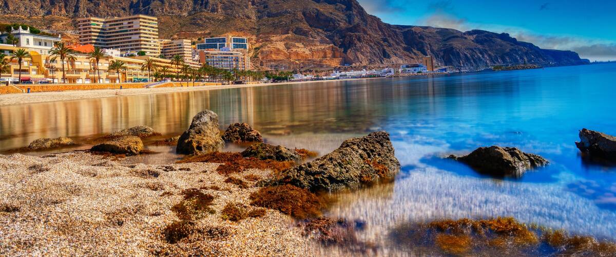 A coastal cityscape with a beach, rocky shoreline, and mountains in the background.