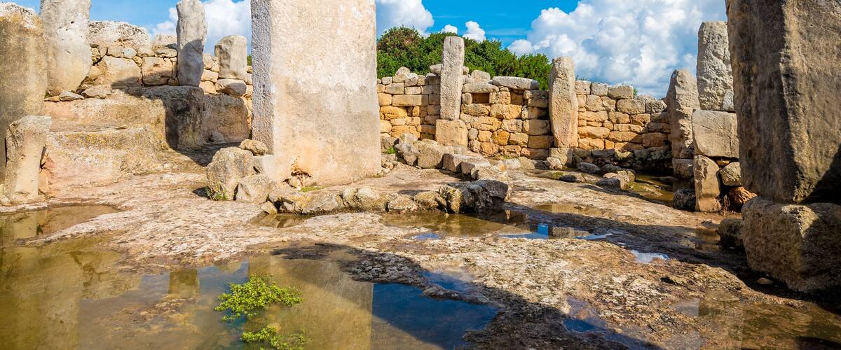 Ancient stone ruins with a large standing stone in the center, surrounded by smaller stones and a clear blue sky.
