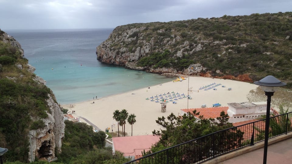 A coastal beach with clear turquoise waters, a rocky cliff, and a row of beach umbrellas.