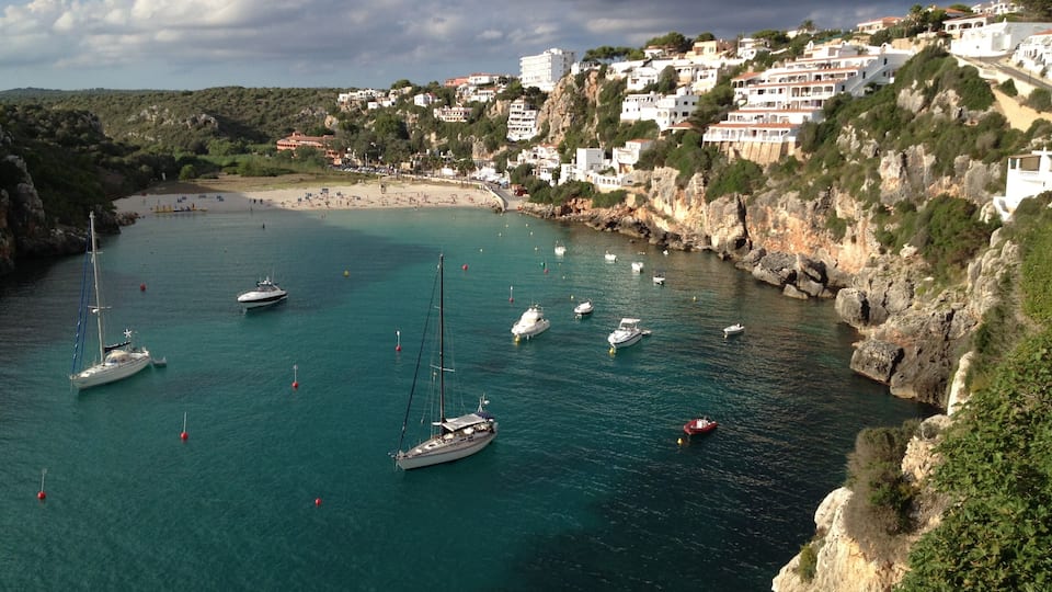 A coastal town with white buildings, a rocky shoreline, and several boats anchored in the water.