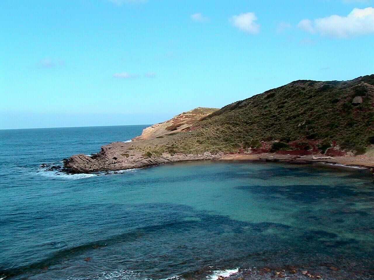 A coastal landscape with clear blue waters, rocky outcrops, and a hill with sparse vegetation.