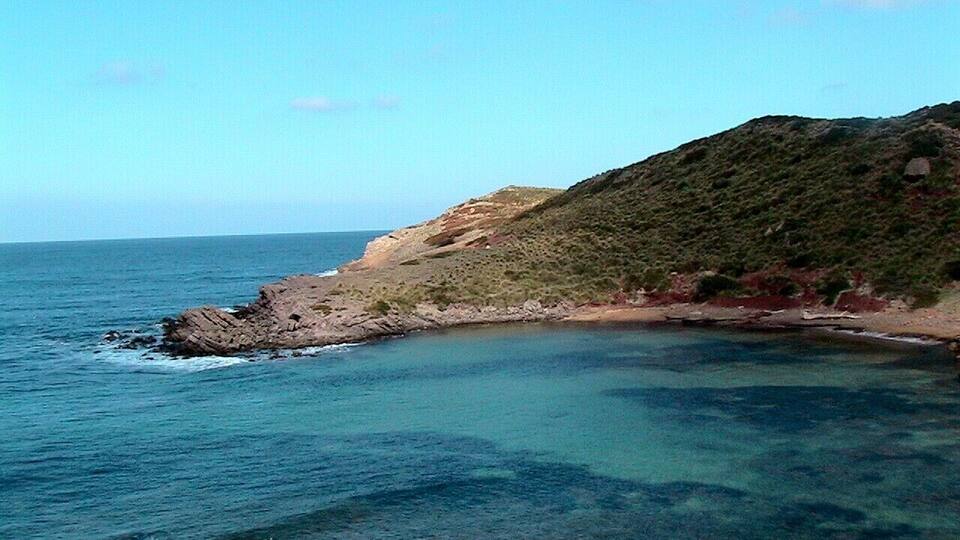 A coastal landscape with clear blue waters, rocky outcrops, and a hill with sparse vegetation.